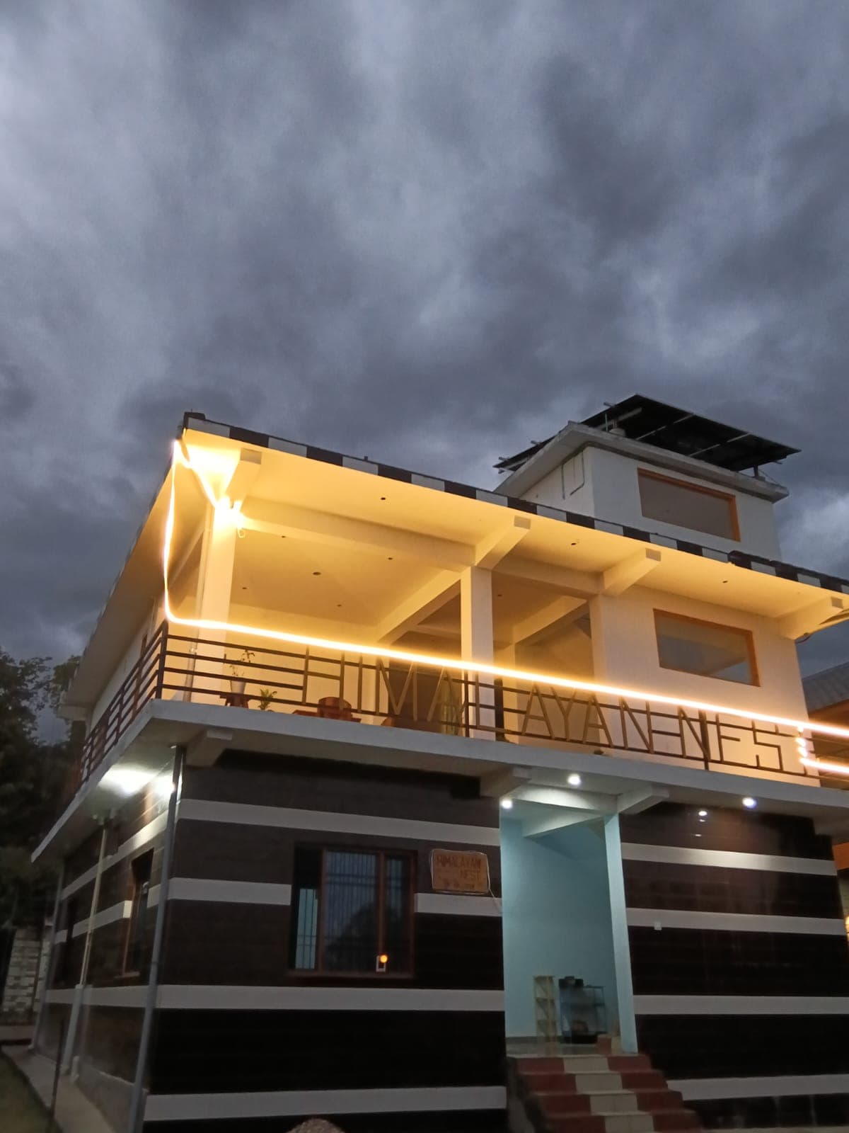 The lit-up balcony of Himalayan Nest under a stormy monsoon sky, with rooftop solar panels.